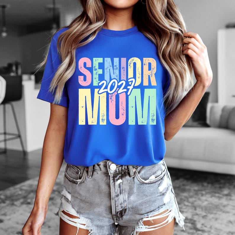 Puede incluir: Una camiseta azul real con las palabras "SENIOR 2023 MOM" en colores pastel. El texto est&aacute; centrado en la camiseta. La camiseta se lleva con pantalones cortos vaqueros.