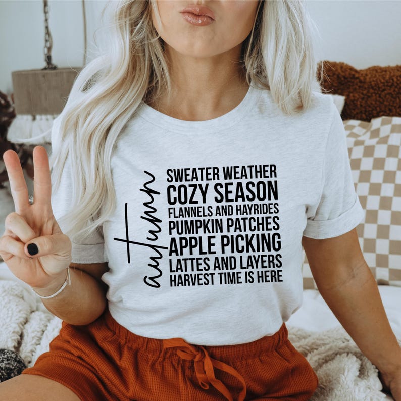 K&ouml;nnte beinhalten: Ein hellgraues T-Shirt mit dem Wort "Autumn" in Schreibschrift und einer Liste von herbstlichen Phrasen in Schwarz. Die Phrasen umfassen "Sweater Weather", "Cozy Season" und "Apple Picking".