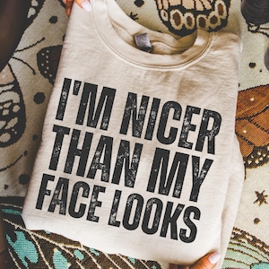 Pu&ograve; includere: Una felpa beige con la scritta "I'M NICER THAN MY FACE LOOKS" in lettere nere sbiadite. La felpa &egrave; piegata e appoggiata su una superficie a fantasia. L'immagine &egrave; un primo piano.