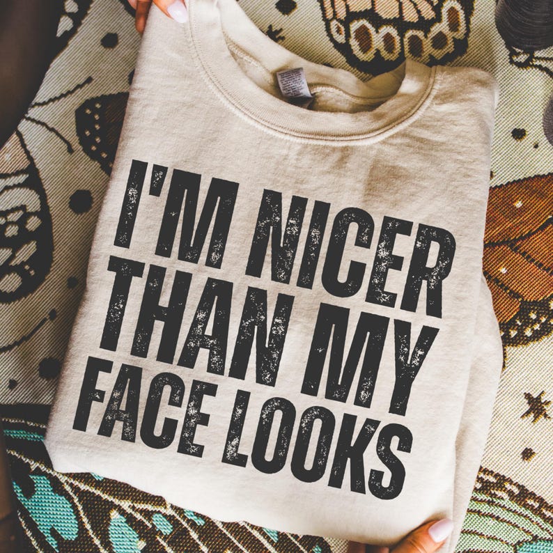 Pu&ograve; includere: Una felpa beige con la scritta "I'M NICER THAN MY FACE LOOKS" in lettere nere sbiadite. La felpa &egrave; piegata e appoggiata su una superficie a fantasia. L'immagine &egrave; un primo piano.