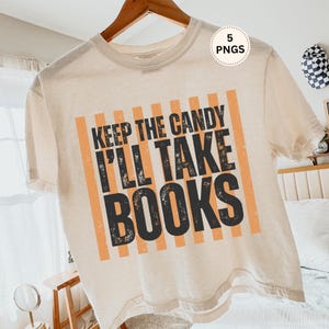 May include: Cream-colored t-shirt with the text "KEEP THE CANDY I'LL TAKE BOOKS" in black and orange. The design features vertical orange stripes. The shirt is hanging on a wooden hanger.