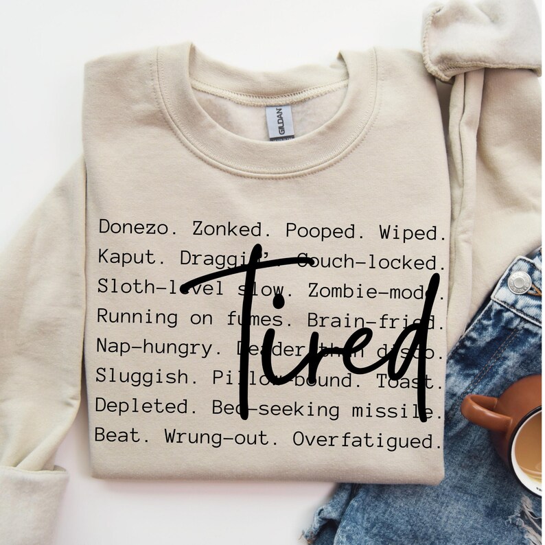 May include: A beige sweatshirt with the text "Donezo. Zonked. Pooped. Wiped. Kaput. Draggi. Couch-locked. Sloth-level slow. Zombie-mod. Running on fumes. Brain-fried. Nap-hungry. Leader. Tired. Sluggish. Pillow bound. Toast. Depleted. Bed-seeking missile. Beat. Wrung-out. Overfatigued." printed on the front.