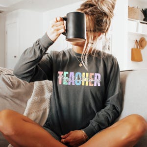 K&ouml;nnte beinhalten: Dunkelgraues Langarmshirt mit dem Wort "TEACHER" in bunten Pastellbuchstaben. Der Name "Mrs. Williams" ist in einer Schreibschrift unter dem Wort "TEACHER" geschrieben. Eine schwarze Tasse wird vor dem Gesicht gehalten.
