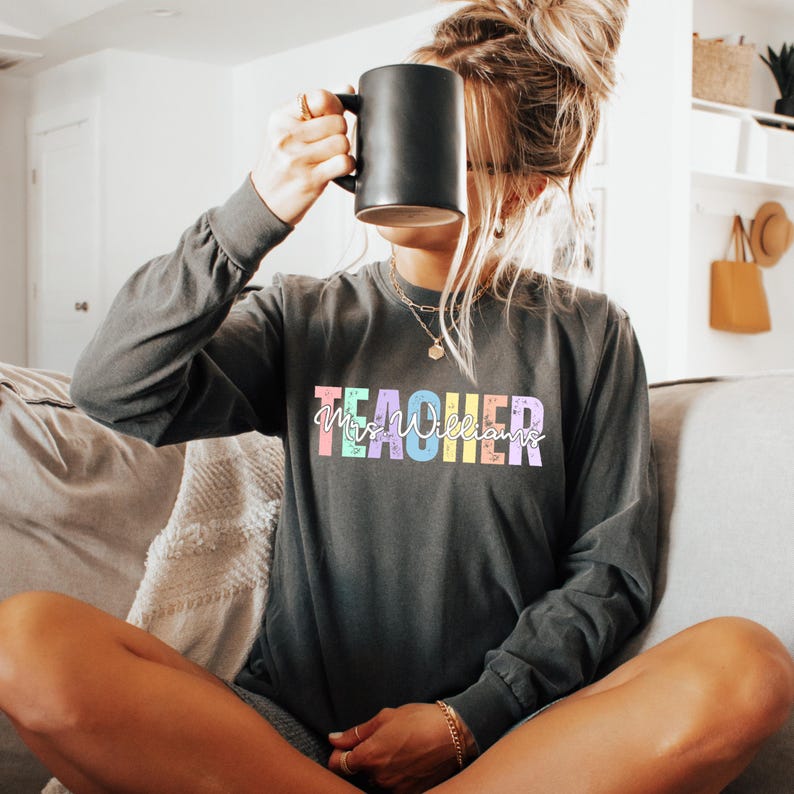 K&ouml;nnte beinhalten: Dunkelgraues Langarmshirt mit dem Wort "TEACHER" in bunten Pastellbuchstaben. Der Name "Mrs. Williams" ist in einer Schreibschrift unter dem Wort "TEACHER" geschrieben. Eine schwarze Tasse wird vor dem Gesicht gehalten.