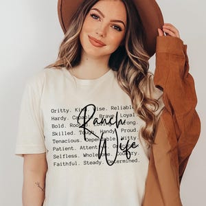 May include: A cream-colored short-sleeve t-shirt featuring a list of black printed adjectives including Gritty, Kind, Wise, Reliable, Hardy, Capable, Brave, Loyal, Bold, Rooted, Loving, Strong, Skilled, Tough, Handy, Proud, Tenacious, Dependable, Witty, Patient, Attentive, Gracious, Selfless, Wholesome, Country, Faithful, Steady, Determined, with the words Ranch Wife written in a large black script font over the text.