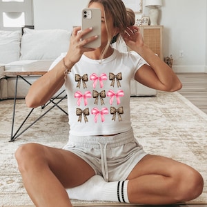 Puede incluir: Una camiseta blanca de manga corta con un patr&oacute;n de cuadr&iacute;cula repetitivo de nueve lazos. El dise&ntilde;o alterna entre lazos de color rosa s&oacute;lido y lazos con estampado de leopardo marr&oacute;n, dispuestos en tres filas de tres en la parte frontal.