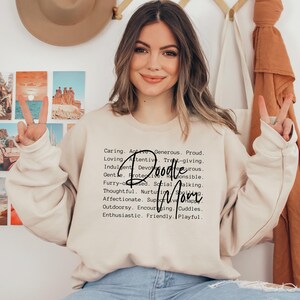 May include: A light beige sweatshirt with black text that reads "Caring. Active. Generous. Proud. Loving. Attentive. Treat-giving. Indulgent. Devourous. Gentle. Protective. Responsible. Doodle Mom. Furry-obsessed. Social. Walking. Thoughtful. Nurturing. Affectionate. Supportive. Happy. Outdoorsy. Encouraging. Cuddles. Enthusiastic. Friendly. Playful."