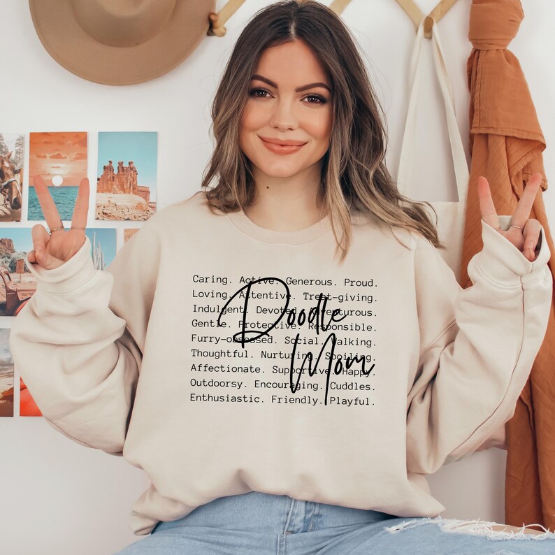 May include: A light beige sweatshirt with black text that reads "Caring. Active. Generous. Proud. Loving. Attentive. Treat-giving. Indulgent. Devourous. Gentle. Protective. Responsible. Doodle Mom. Furry-obsessed. Social. Walking. Thoughtful. Nurturing. Affectionate. Supportive. Happy. Outdoorsy. Encouraging. Cuddles. Enthusiastic. Friendly. Playful."