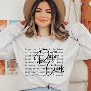 May include: A light gray crewneck sweatshirt featuring black text that lists professional traits including Organized, Efficient, Reliable, Structured, Logical, Numerical, Database-driven, Communicative, Detail-oriented, Team-oriented, Meticulous, Thorough, Diligent, Friendly, Accurate, Tech-savvy, Resourceful, Proactive, Data-savvy, Analytical, Managerial, Helpful, Trustworthy, Discreet, with the words Data Clerk written in a large black script font over the text.
