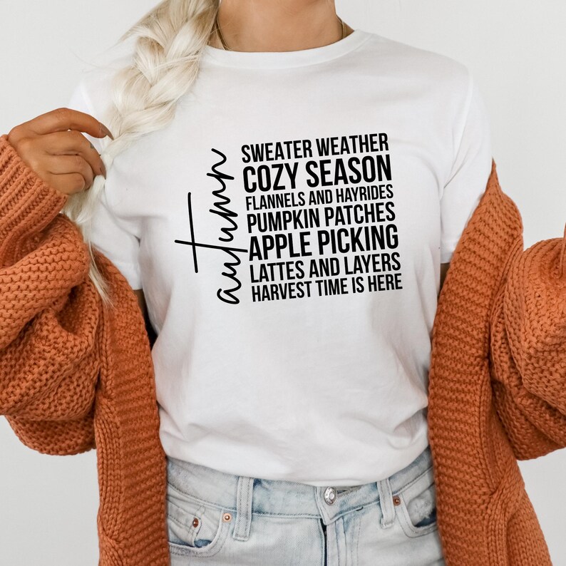 K&ouml;nnte beinhalten: Ein wei&szlig;es T-Shirt mit dem Wort "Herbst" in Schreibschrift und einer Liste von herbstlichen Phrasen in Schwarz, darunter "Sweater Weather" und "Apple Picking". Ein orangefarbener Cardigan ist &uuml;ber den Schultern drapiert.