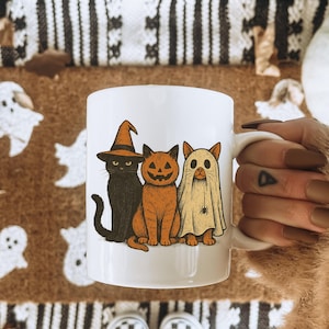 K&ouml;nnte beinhalten: Wei&szlig;e Keramiktasse mit drei Halloween-Katzen. Eine ist schwarz und tr&auml;gt einen Hexenhut, die zweite ist orange mit einem K&uuml;rbisgesicht und die dritte ist ein Geist. Die Tasse hat einen wei&szlig;en Henkel.