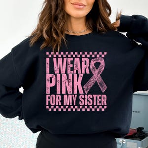 May include: Black sweatshirt with the text "I WEAR PINK FOR MY SISTER" in pink, along with a pink ribbon graphic. The text is framed by a pink and white checkered pattern.