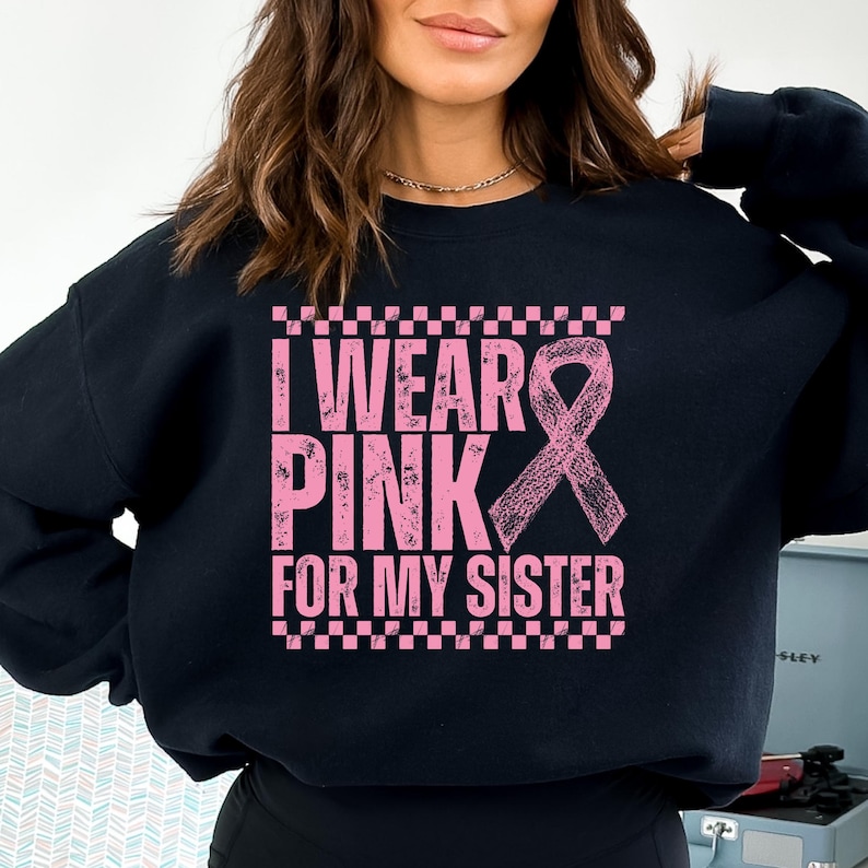 May include: Black sweatshirt with the text "I WEAR PINK FOR MY SISTER" in pink, along with a pink ribbon graphic. The text is framed by a pink and white checkered pattern.