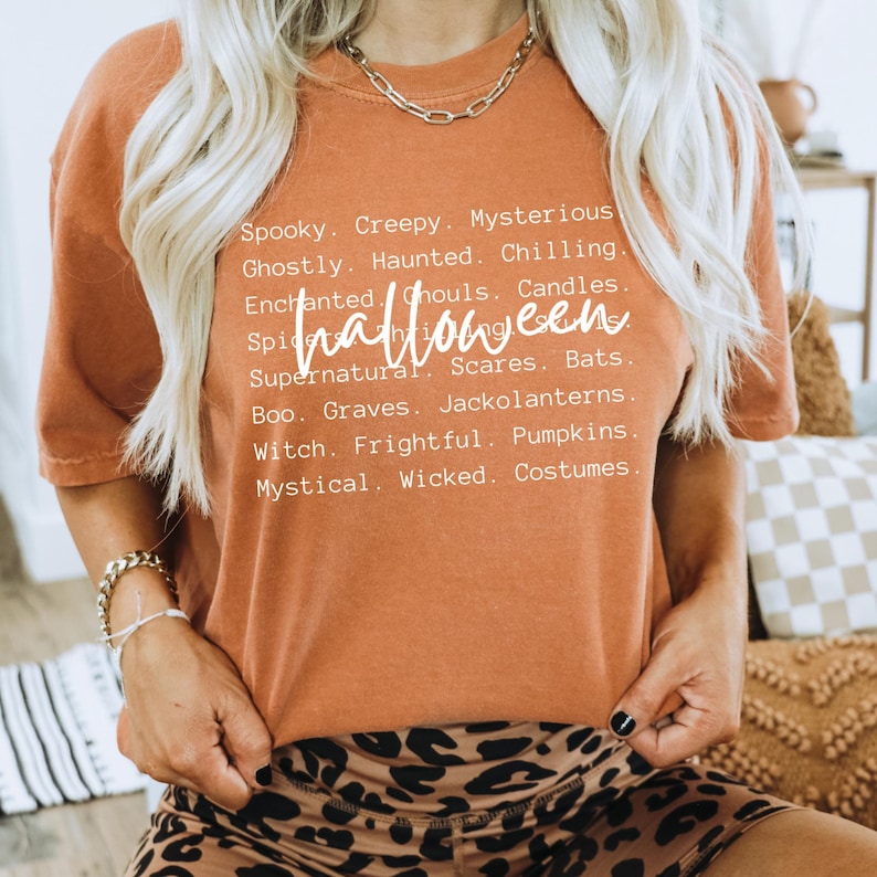 May include: An orange t-shirt with the word "Halloween" in a cursive font. The shirt features a list of Halloween-themed words in white, including "Spooky," "Creepy," and "Mysterious." The model is wearing a gold chain necklace and a gold bracelet.