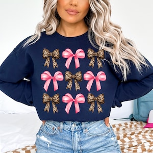 Puede incluir: Una sudadera azul marino de cuello redondo con un patr&oacute;n repetitivo de nueve lazos. El dise&ntilde;o alterna entre lazos de color rosa claro s&oacute;lido y lazos con estampado de leopardo marr&oacute;n y negro, dispuestos en tres filas de tres en el pecho.