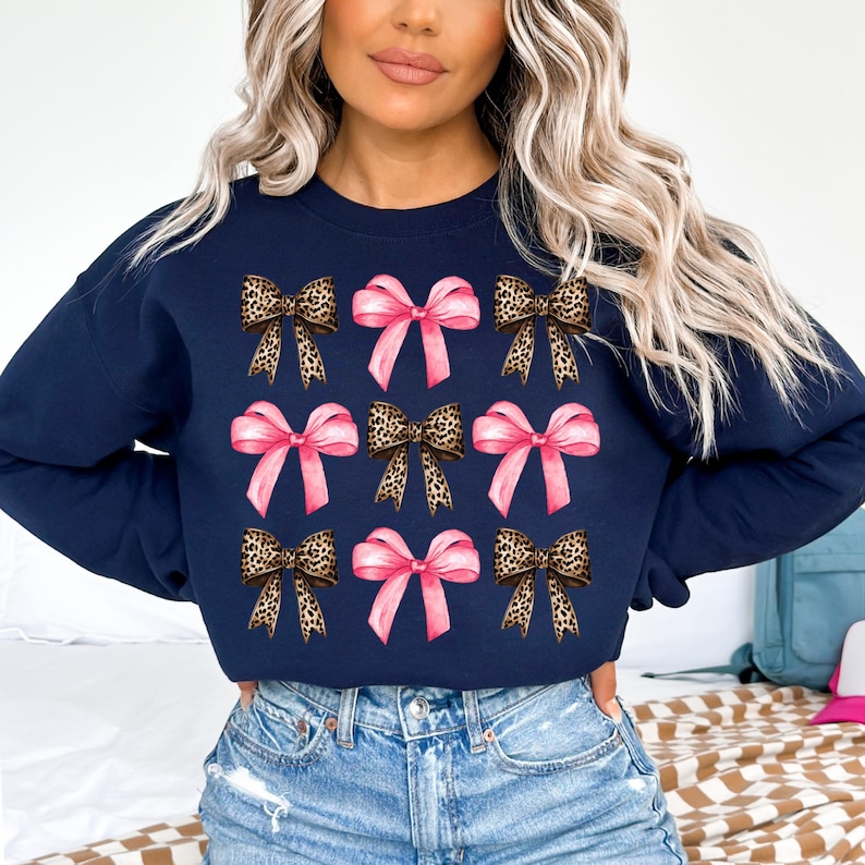 Puede incluir: Una sudadera azul marino de cuello redondo con un patr&oacute;n repetitivo de nueve lazos. El dise&ntilde;o alterna entre lazos de color rosa claro s&oacute;lido y lazos con estampado de leopardo marr&oacute;n y negro, dispuestos en tres filas de tres en el pecho.
