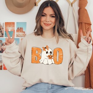 以下が含まれることがあります： オレンジ色の文字で「BOO」と書いてあり、クモの巣模様の入った、かわいい幽霊がカボチャを持っているベージュのトレーナーです。