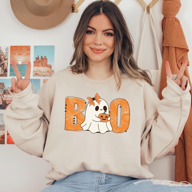 以下が含まれることがあります： オレンジ色の文字で「BOO」と書いてあり、クモの巣模様の入った、かわいい幽霊がカボチャを持っているベージュのトレーナーです。