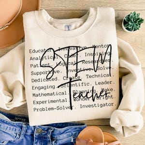 May include: A cream-colored crewneck sweatshirt featuring black text that lists various qualities like Educator, Analytical, Patient, and Problem-Solver, with the large black script text STEM Teacher overlaid across the center of the words.