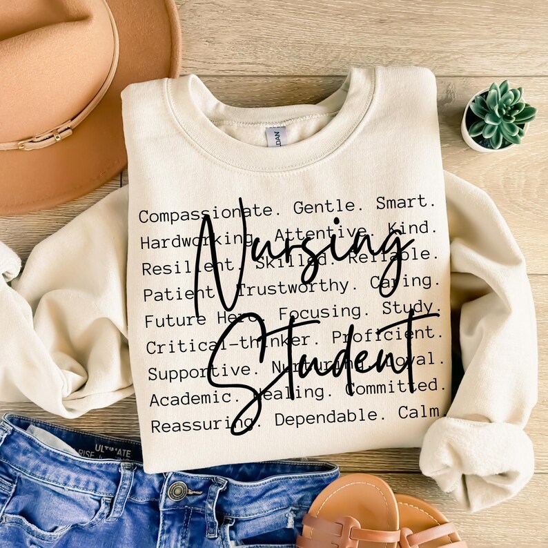 May include: A beige sweatshirt with the text "Nursing Student" in black lettering. The text is surrounded by a list of positive adjectives describing a nursing student, such as compassionate, gentle, smart, hardworking, attentive, kind, resilient, skilled, reliable, patient, trustworthy, caring, future hero, focusing, study, critical-thinker, proficient, supportive, nurturing, loyal, academic, healing, committed, reassuring, dependable, and calm.