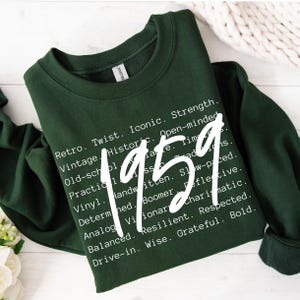 May include: A dark green sweatshirt with the year "1959" in large white text. The sweatshirt also features a list of words like "Retro," "Iconic," and "Resilient" in white text. The Gildan brand tag is visible.