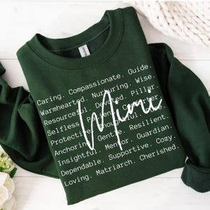 May include: A dark green sweatshirt with white text that reads "Caring. Compassionate. Guide. Warmhearted. Nurturing. Wise. Resourceful. Devoted. Pillar. Mimi. Selfless. Patient. Carter. Protective. Thoughtful. Anchoring. Gentle. Resilient. Insightful. Mentor. Guardian. Dependable. Supportive. Cozy. Loving. Matriarch. Cherished."