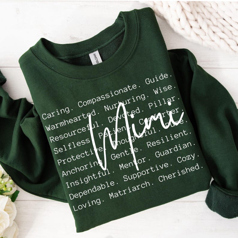 May include: A dark green sweatshirt with white text that reads "Caring. Compassionate. Guide. Warmhearted. Nurturing. Wise. Resourceful. Devoted. Pillar. Mimi. Selfless. Patient. Carter. Protective. Thoughtful. Anchoring. Gentle. Resilient. Insightful. Mentor. Guardian. Dependable. Supportive. Cozy. Loving. Matriarch. Cherished."