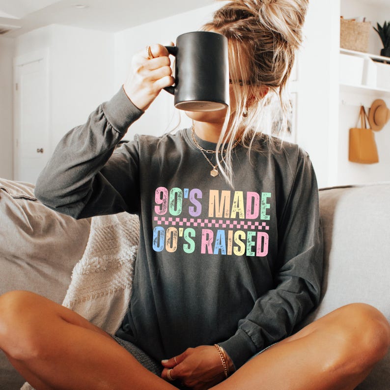 May include: Dark grey long-sleeve shirt with the text "90'S MADE 00'S RAISED" in pastel colors. The shirt has a crew neck and is made of a soft material. A black mug is held up to the face.