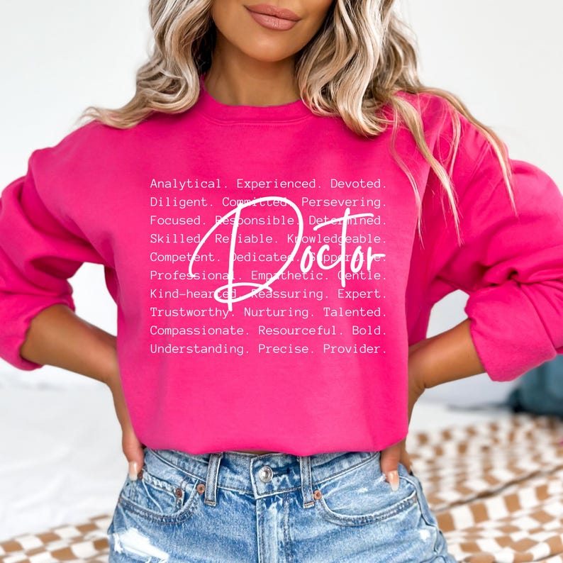 May include: A pink sweatshirt with white text that reads "Analytical. Experienced. Devoted. Diligent. Committed. Persevering. Focused. Responsible. Determined. Skilled. Reliable. Knowledgeable. Competent. Dedicate. Doctor. Professional. Empathetic. Gentle. Kind-hearted. Reassuring. Expert. Trustworthy. Nurturing. Talented. Compassionate. Resourceful. Bold. Understanding. Precise. Provider."
