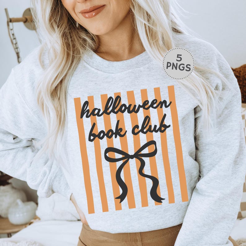 May include: A light gray sweatshirt with the text "halloween book club" in black script, set against orange vertical stripes. A black bow graphic is centered below the text. The sweatshirt is a casual, comfortable style.