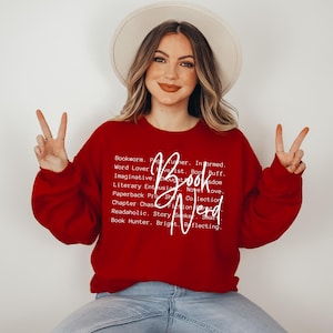 May include: Red sweatshirt with white text that reads "Book Nerd" and a list of words describing a book lover. The model is wearing the sweatshirt and making a peace sign with both hands.