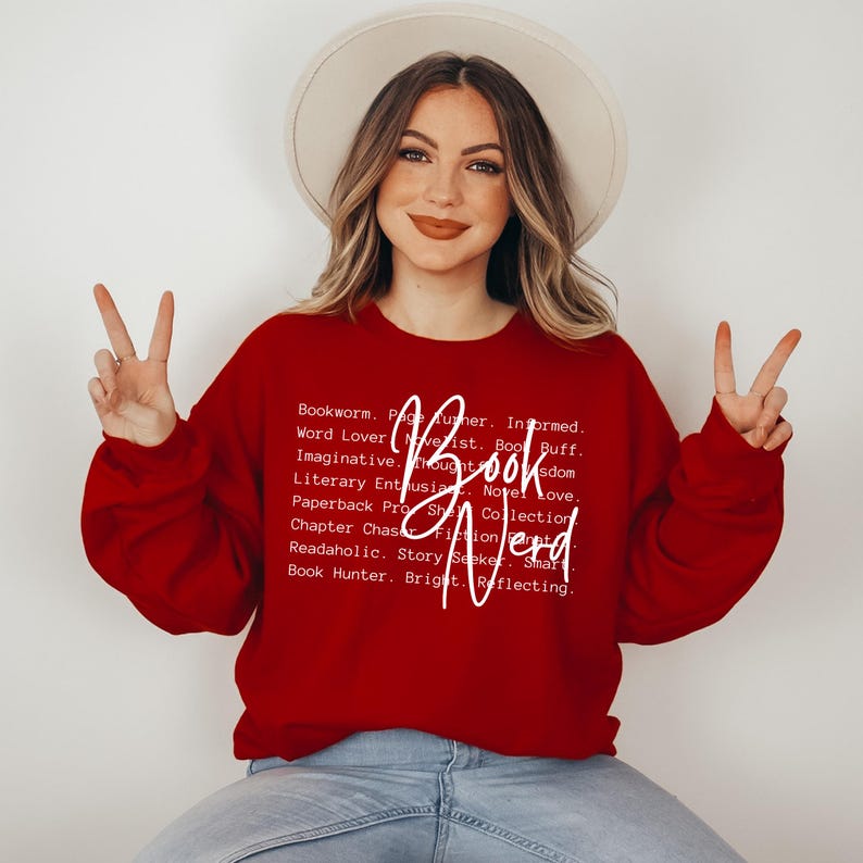 May include: Red sweatshirt with white text that reads "Book Nerd" and a list of words describing a book lover. The model is wearing the sweatshirt and making a peace sign with both hands.