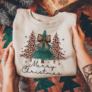May include: A beige crewneck sweatshirt featuring a graphic print of three Christmas trees. The center tree is solid green with a leopard print bow, while the two flanking trees feature a leopard print pattern. The text Merry Christmas is printed below.