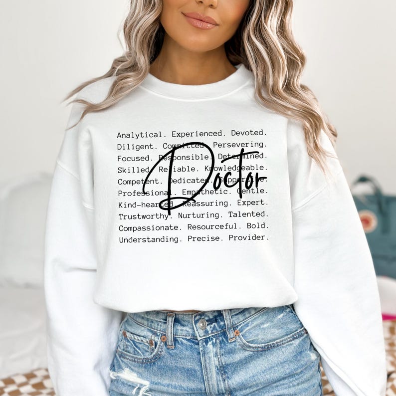 May include: White sweatshirt with the word "Doctor" in black text and a list of positive attributes describing a doctor, such as analytical, experienced, devoted, diligent, committed, persevering, focused, responsible, determined, skilled, reliable, knowledgeable, competent, dedicated, professional, empathetic, gentle, kind-hearted, reassuring, expert, trustworthy, nurturing, talented, compassionate, resourceful, bold, understanding, precise, and provider.