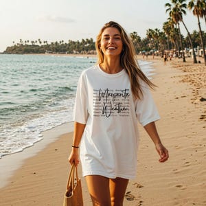May include: A white t-shirt with the words "Margarita Weather" printed on it. The shirt is worn by a person walking on a sandy beach. The text includes words like "Tequila", "Salty", "Frozen", "Cocktail", "Sipping", "Cheers", and "Happy Hour".