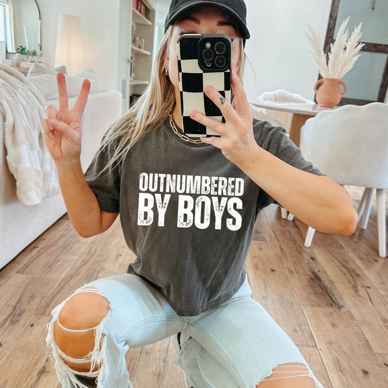 May include: Dark gray t-shirt with the text "OUTNUMBERED BY BOYS" in white block letters. The shirt is worn with light blue jeans and a black baseball cap. A gold chain necklace is visible.