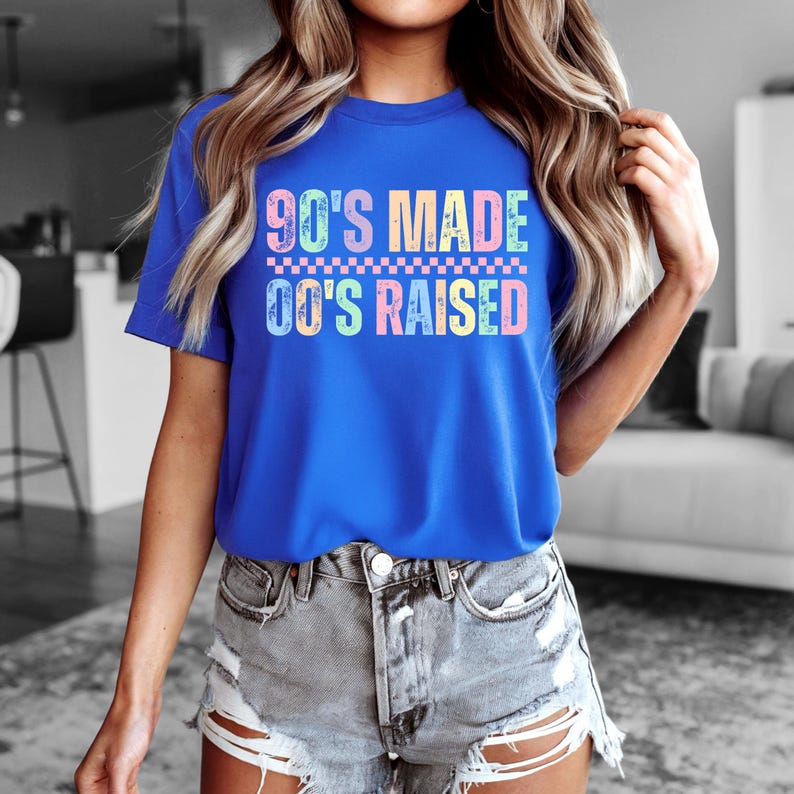 May include: A royal blue t-shirt with the text "90'S MADE 00'S RAISED" in colorful, retro-style lettering. The shirt has a crew neck and short sleeves. The design includes a checkered pattern between the two phrases.