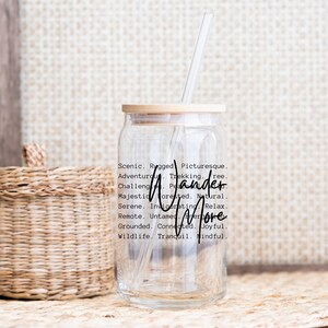 K&ouml;nnte beinhalten: Ein klares Trinkglas mit Bambusdeckel und einem durchsichtigen Strohhalm. Auf dem Glas stehen die Worte "Wander More" in schwarzer Schrift. Um die Worte herum stehen beschreibende W&ouml;rter wie "Scenic", "Rugged" und "Adventurous".
