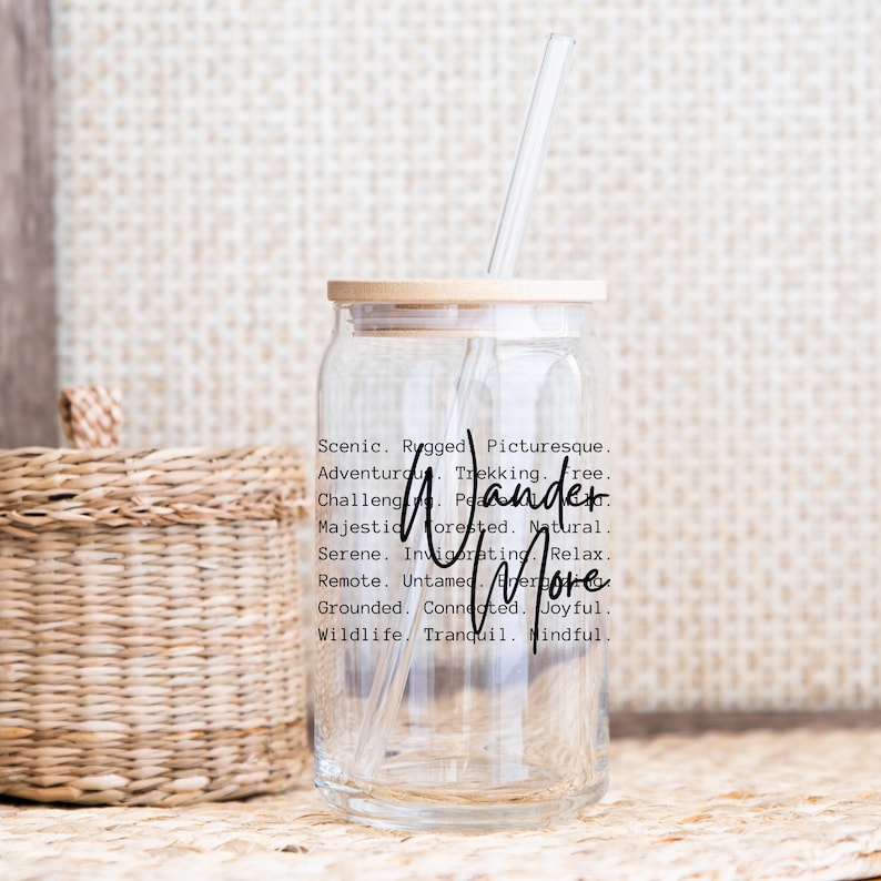 K&ouml;nnte beinhalten: Ein klares Trinkglas mit Bambusdeckel und einem durchsichtigen Strohhalm. Auf dem Glas stehen die Worte "Wander More" in schwarzer Schrift. Um die Worte herum stehen beschreibende W&ouml;rter wie "Scenic", "Rugged" und "Adventurous".