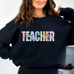 K&ouml;nnte beinhalten: Schwarzes Sweatshirt mit dem Wort "TEACHER" in pastellfarbenen Blockbuchstaben &uuml;ber "Mrs. Hill" in einer Schreibschrift. Das Sweatshirt hat einen Rundhalsausschnitt.
