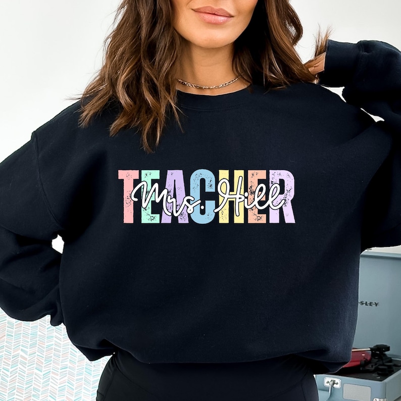 K&ouml;nnte beinhalten: Schwarzes Sweatshirt mit dem Wort "TEACHER" in pastellfarbenen Blockbuchstaben &uuml;ber "Mrs. Hill" in einer Schreibschrift. Das Sweatshirt hat einen Rundhalsausschnitt.