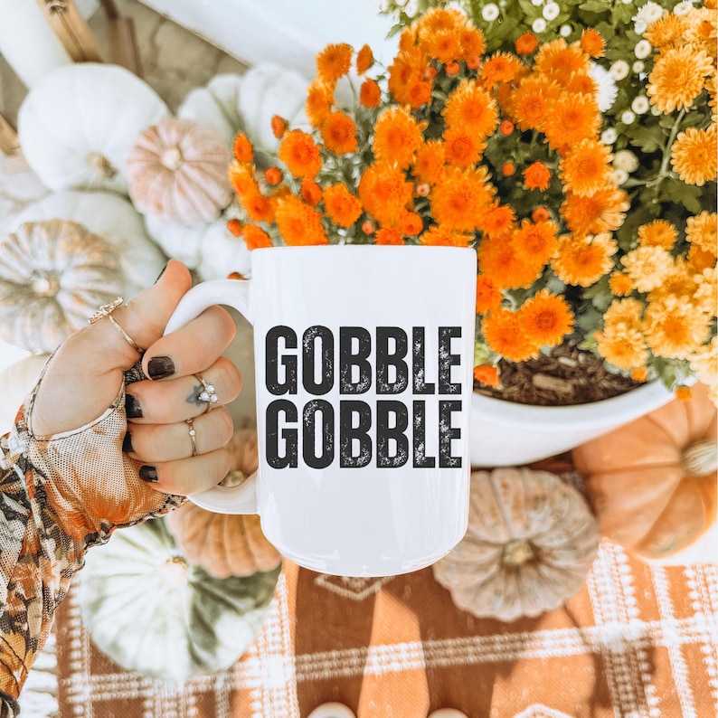 May include: White ceramic mug with the words "GOBBLE GOBBLE" in black, held in a hand with rings. The mug is surrounded by pumpkins, orange and yellow flowers, and a patterned surface, evoking a fall theme.