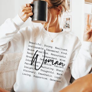 May include: A white long-sleeve sweatshirt featuring a list of black printed adjectives including Beautiful, Strong, Resilient, Unstoppable, Empowered, Kind, Brave, Fearless, Resourceful, Tenacious, Independent, Wise, Grounded, Determined, Intelligent, Gentle, Patient, Perceptive, Nurturing, Sharp, Insightful, Thoughtful, Warm, Visionary, Empathetic, Loyal, Loving, Supportive, Generous, with the word Woman written in a large black script font across the center.