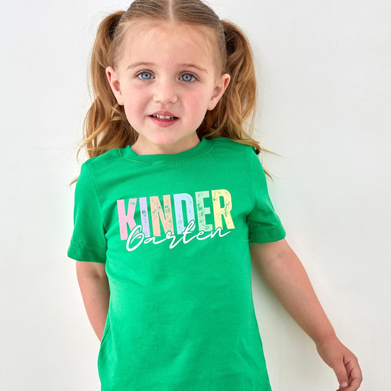 K&ouml;nnte beinhalten: Ein Kind tr&auml;gt ein leuchtend gr&uuml;nes T-Shirt mit dem Aufdruck "KINDER GARTEN" in Pastellfarben. Das Kind hat blondes Haar und blaue Augen und l&auml;chelt in die Kamera.