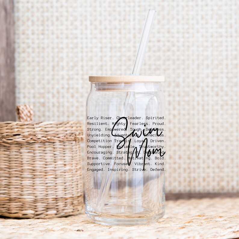 May include: A clear glass tumbler with a wooden lid and a straw. The tumbler has a black and white design with the words "Swim Mom" and a list of positive adjectives describing a strong woman.