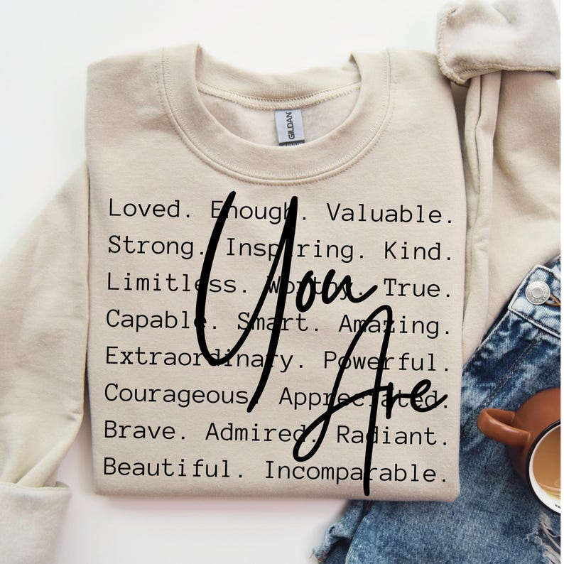 May include: A beige sweatshirt with the text "You Are" written in black cursive letters. The text is surrounded by a list of positive adjectives, including "Loved", "Enough", "Valuable", "Strong", "Inspiring", "Kind", "Limitless", "True", "Capable", "Smart", "Amazing", "Extraordinary", "Powerful", "Courageous", "Appreciated", "Brave", "Admired", "Radiant", "Beautiful", and "Incomparable".