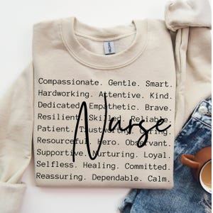 K&ouml;nnte beinhalten: Beigefarbener Pullover mit dem Wort "Nurse" in gro&szlig;en schwarzen Buchstaben, umgeben von einer Liste positiver Adjektive, die die Eigenschaften einer Krankenschwester beschreiben.