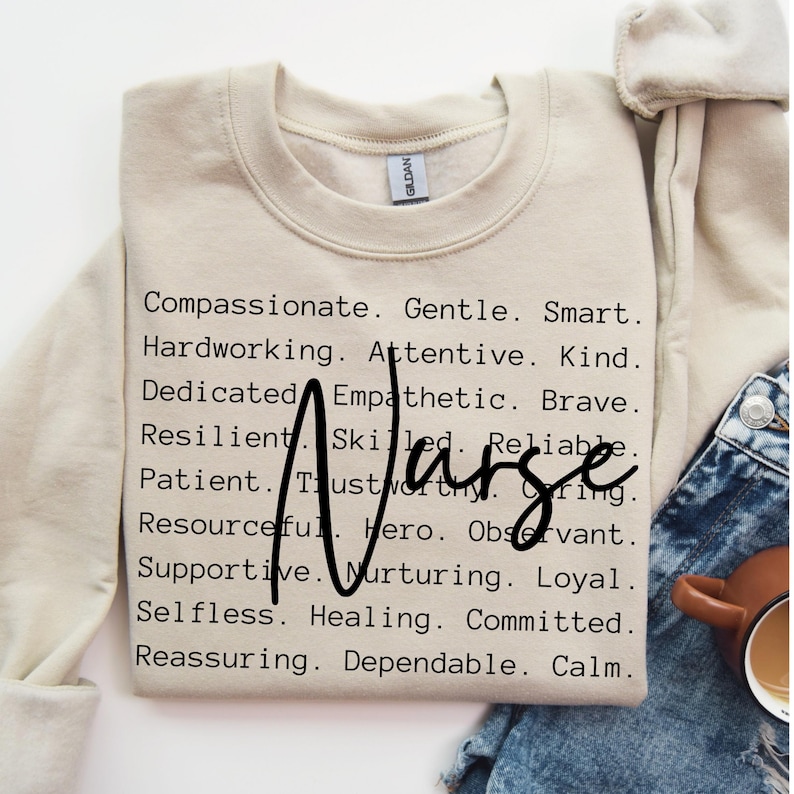 K&ouml;nnte beinhalten: Beigefarbener Pullover mit dem Wort "Nurse" in gro&szlig;en schwarzen Buchstaben, umgeben von einer Liste positiver Adjektive, die die Eigenschaften einer Krankenschwester beschreiben.