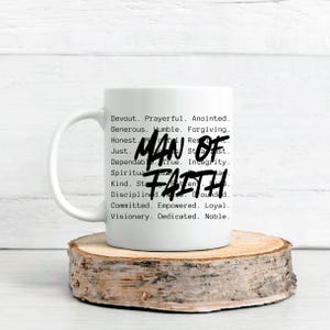 Op de afbeelding: Witte keramische mok met de woorden "MAN OF FAITH" in zwart script. De mok heeft ook een lijst met positieve eigenschappen in een zwarte, typemachine-achtige letter. De mok wordt gepresenteerd op een houten onderzetter.