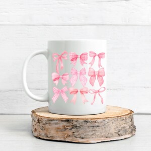 Puede incluir: Taza de cer&aacute;mica blanca con una colecci&oacute;n de lazos rosas de acuarela en varios estilos. Los lazos incluyen dise&ntilde;os lisos, de cuadros y de cinta. La taza se exhibe sobre una rodaja de madera.